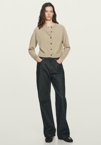 Vrouw staat met handen in de zakken, draagt een beige gebreid vest met knopen, losse donkere jeans en zwarte schoenen tegen een effen lichte achtergrond.