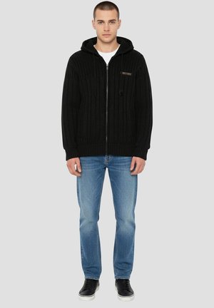 Sort strikket hættejakke med lynlås, ribsstruktur, justerbare snore og logopatch, kombineret med lyseblå jeans og sorte sneakers.