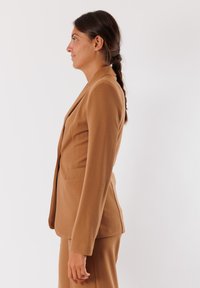 Blazer marrone su misura realizzato in tessuto morbido, caratterizzato da una vestibilità slim, revers a punte e tasche laterali, mostrato da una vista laterale.