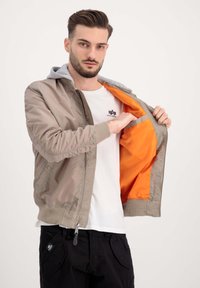 Kerge pommitaja jope taupe värviga halli kapuutsiga. Omab oranži voodrit, soonikuga varrukaotsasid, lukuga kinni ja küljetaskuid.