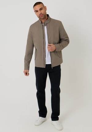 Mand iført taupe lynlåstrøje, hvid t-shirt, sorte bukser og hvide sneakers mod en ensfarvet baggrund.