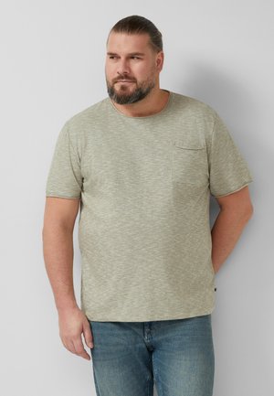 Bärtiger Mann mit zurückgebundenem Haar, trägt ein hellgraues kurzärmeliges Hemd mit Tasche und blaue Jeans, steht vor einer schlichten hellen Wand.