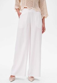 Wijdvallende witte broek gemaakt van soepel materiaal, met steekzakken en een hoge tailleband. Gecombineerd met een gestructureerd beige gebreid topje en roze schoenen.