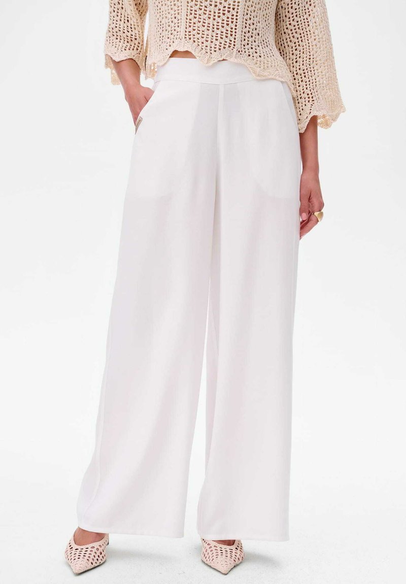 Wijdvallende witte broek gemaakt van soepel materiaal, met steekzakken en een hoge tailleband. Gecombineerd met een gestructureerd beige gebreid topje en roze schoenen.