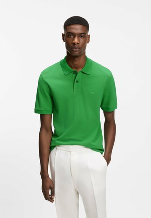 Mand iført en knaldrød grøn kortærmet polo-shirt med hvide bukser, stående med den ene hånd i lommen mod en ensfarvet baggrund.
