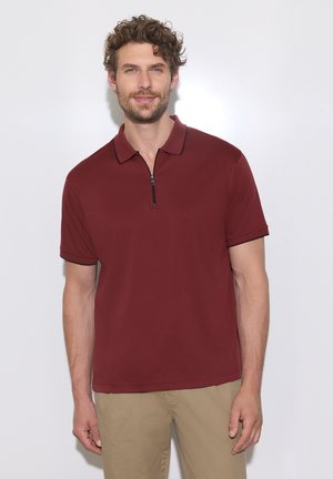 Mann mit lockigem Haar, der ein kurzärmeliges burgunderfarbenes Poloshirt mit Reißverschluss vorne und beigen Hosen trägt und vor einem weißen Hintergrund steht.