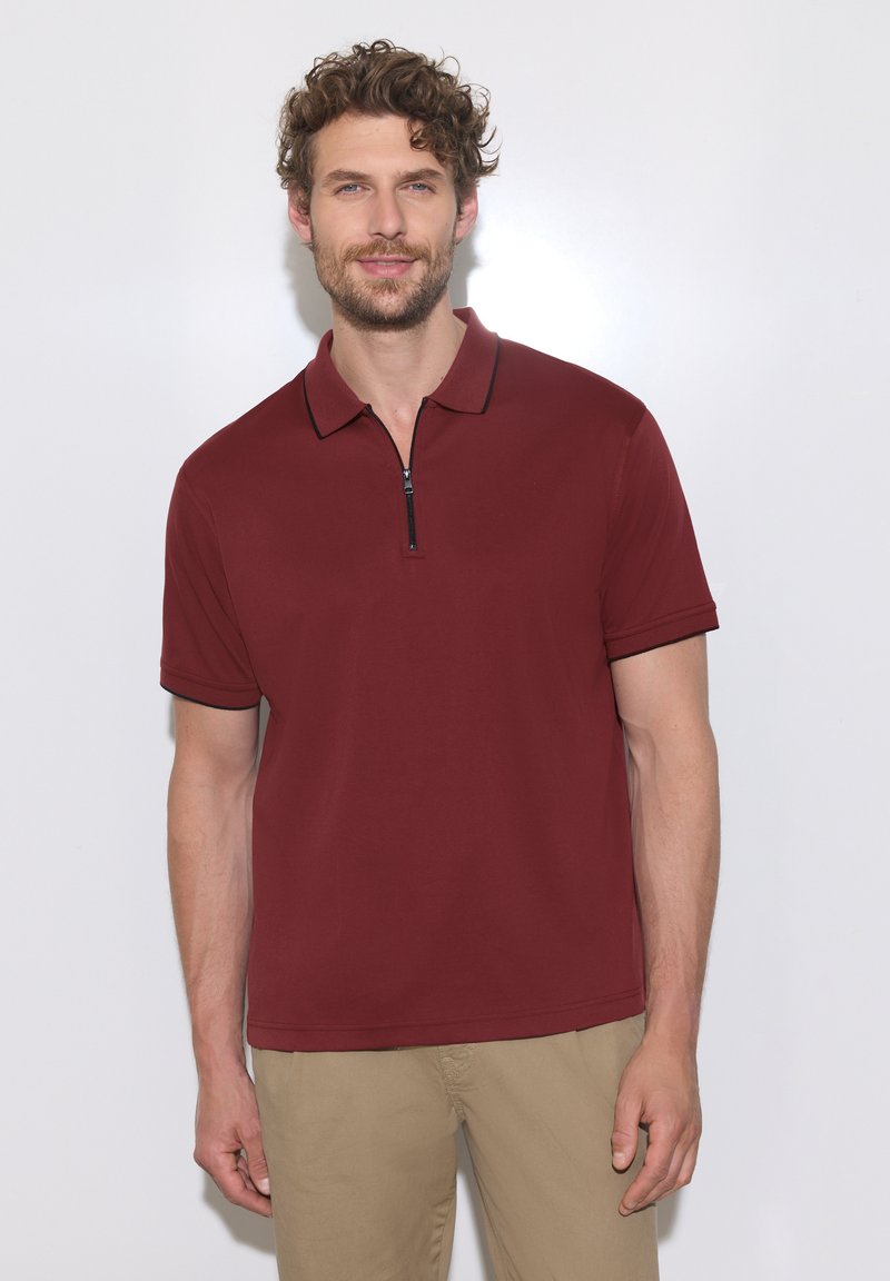 Homme aux cheveux bouclés portant un polo grenat à manches courtes avec une fermeture éclair à l'avant et un pantalon beige, debout devant un fond blanc.