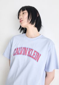 Helles blaues Baumwoll-T-Shirt mit dem Text "CALVIN KLEIN" in fetter roter Schrift. Kurze Ärmel und lockere Passform. Einfarbiger Hintergrund.