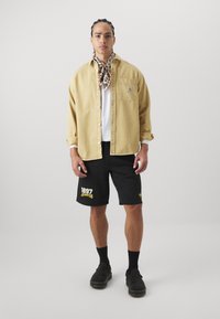 Tan button-up shirt over a white t-shirt, black shorts with yellow text, and black shoes. Accessorized with a patterned scarf.