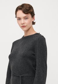 Mørkegrå, langermet genser med rund hals, teksturert stoff og belte i midjen. Har knappdetaljer på skulderen.
