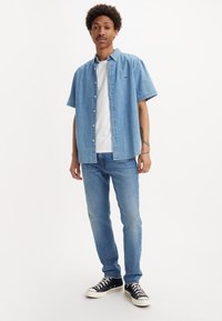 Chemise en denim bleu clair à manches courtes sur un T-shirt blanc, associée à un jean bleu moyen et des sneakers noires avec des semelles blanches.