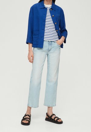 Veste en lin bleue avec une poche avant, t-shirt rayé blanc et bleu, jean large bleu clair, et sandales noires à brides.