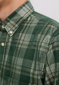 Groen geruit overhemd van corduroy stof, met verticale en horizontale strepen in donkerder groen, bruin en wit, met een button-down kraag.