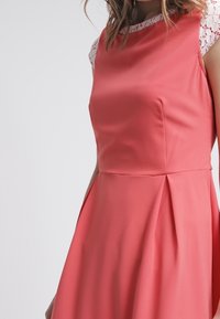 Kobieta ubrana w bezrękawową koralową sukienkę z białymi koronkowymi rękawami, dopasowanym gorsie i rozkloszowanym dołem na jednolitym jasnym tle.