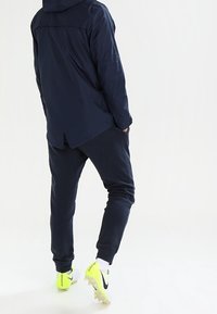 Giacca a vento navy con cappuccio, pantaloni jogger scuri dalla vestibilità rilassata, e scarpini da calcio giallo brillante con dettagli bianchi e suola testurizzata.