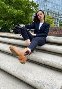 Tan ankle boots with a textured finish and black striped elastic detail. Worn with dark pants and a blazer in an outdoor setting.