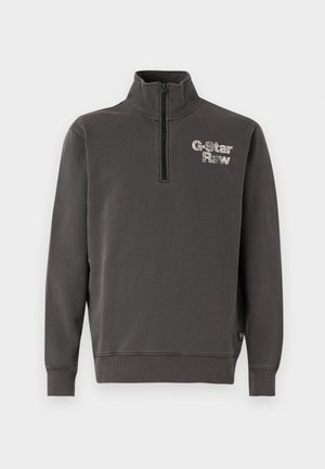 Ciemnoszara bluza zapinana na zamek z wysokim kołnierzem, z jasnoszarym logo po lewej stronie klatki piersiowej oraz prążkowanymi mankietami i dołem. Miękki materiał.