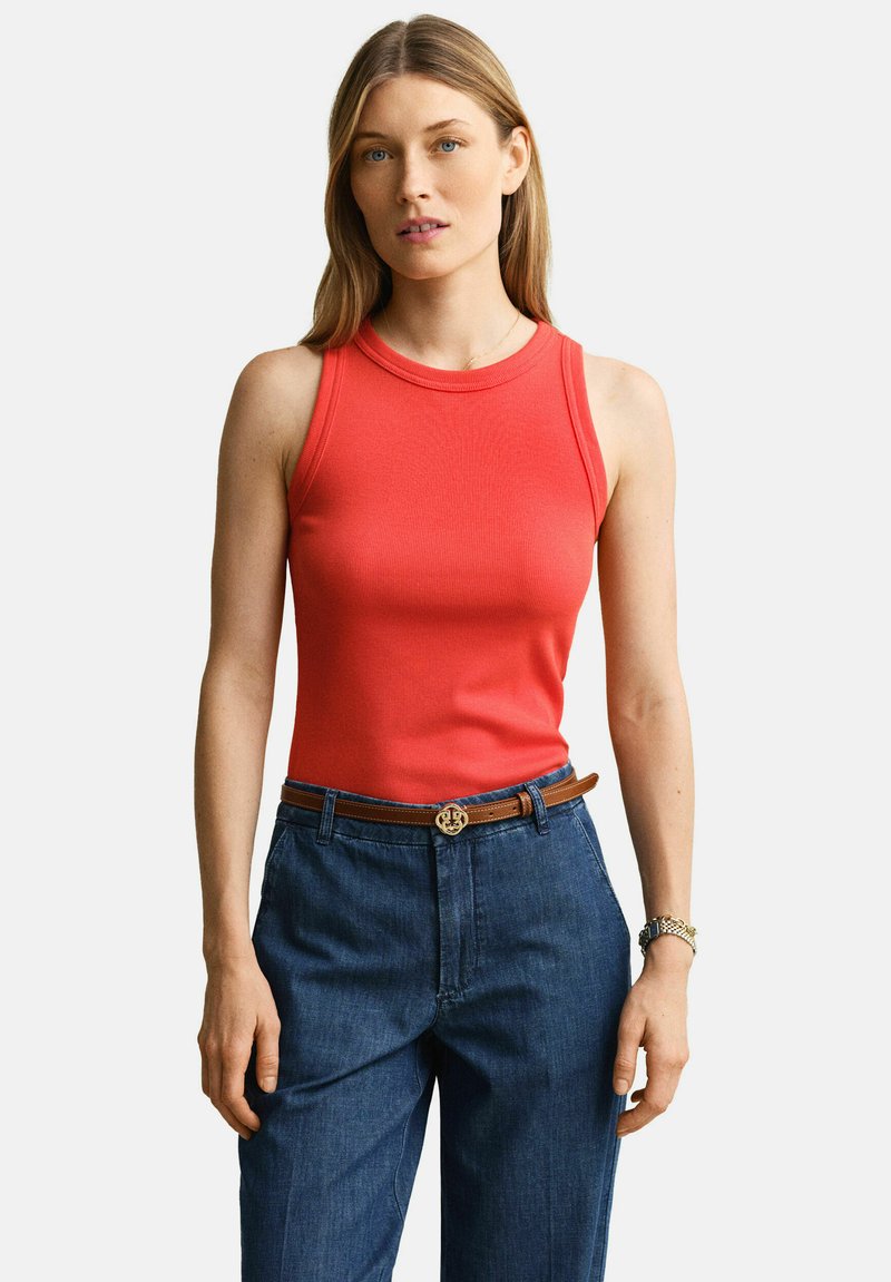 Femme portant un haut rouge sans manches rentré dans un jean bleu taille haute avec une ceinture marron, debout les bras détendus, fond neutre.