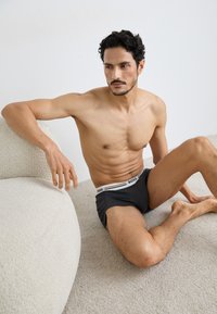 Sivé boxer šortky s bielym pásom s logom "BOSS". Model má krátke tmavé vlasy a svalnatú postavu, sedí na texturovanom koberci.