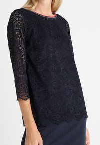 Top in pizzo blu navy con colletto tondo rifinito in rosso, maniche a tre quarti e orlo scallopato. Il tessuto presenta un motivo floreale.