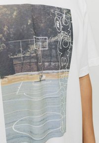 Camiseta blanca de manga corta con un estampado gráfico de una cancha de baloncesto, con un aro y texto al lado en negro y gris.