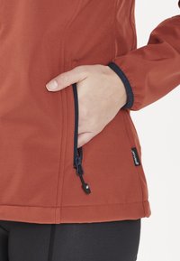 Rostfärgad softshell-jacka med en ficka med dragkedja, elastiska ärmar och en mjuk yta. Svarta accenter vid ärmslutet och fickans dragkedja.