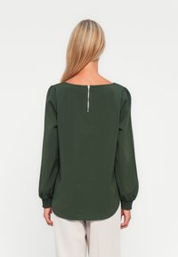 Blusa verde con escote cuadrado, mangas largas abullonadas y puños fruncidos. Presenta un detalle con cremallera en la parte posterior. Textura de tejido suave.