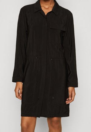 Kobieta w czarnej sukience koszulowej z długim rękawem, z kołnierzykiem, kieszenią z przodu i ściągaczem w talii.