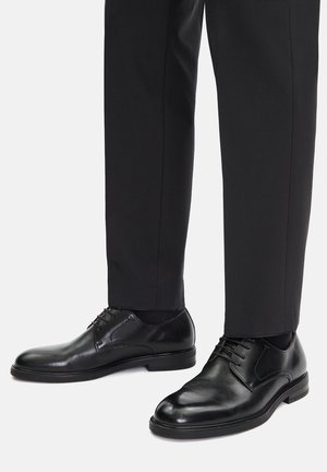 Męskie czarne skórzane eleganckie buty na sznurowadła, noszone z czarnymi eleganckimi spodniami i czarnymi skarpetami na jednolitym jasnym tle.