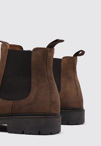 Botas Chelsea de ante marrón con paneles laterales elásticos, bucles para tirar y una suela de goma negra texturizada con un patrón de tacos gruesos.