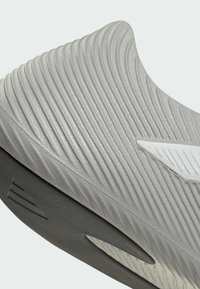 Αθλητικό παπούτσι ανοιχτού γκρι με υφή στο επάνω μέρος, που έχει οριζόντιες ραβδώσεις και λευκή λεπτομέρεια. Μαύρη σόλα με κομψό, αεροδυναμικό σχέδιο.