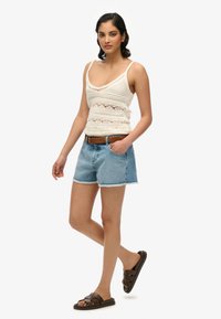 Top de ganchillo blanco sin mangas con escote en V, shorts de mezclilla altos de color azul claro y sandalias de tiras marrones. Textura suave y ajuste casual.