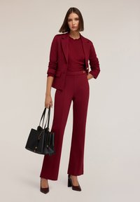 Bordeaux op maat gemaakte pak met lange broek, gestructureerde trui en gestructureerde blazer. Accessoires zijn een zwarte handtas en hoge hakken.