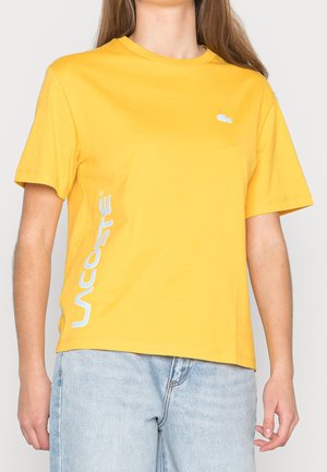 Frau trägt ein gelbes Lacoste-T-Shirt mit weißem Logo auf der Brust und vertikalem Text auf der linken Seite, kombiniert mit hellblauen Jeans.