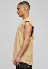Beige mouwloos t-shirt met ruwe randen, zijopeningen, ontspannen pasvorm. Draagt bij donkere textuur broek, gepresenteerd tegen een neutrale achtergrond.
