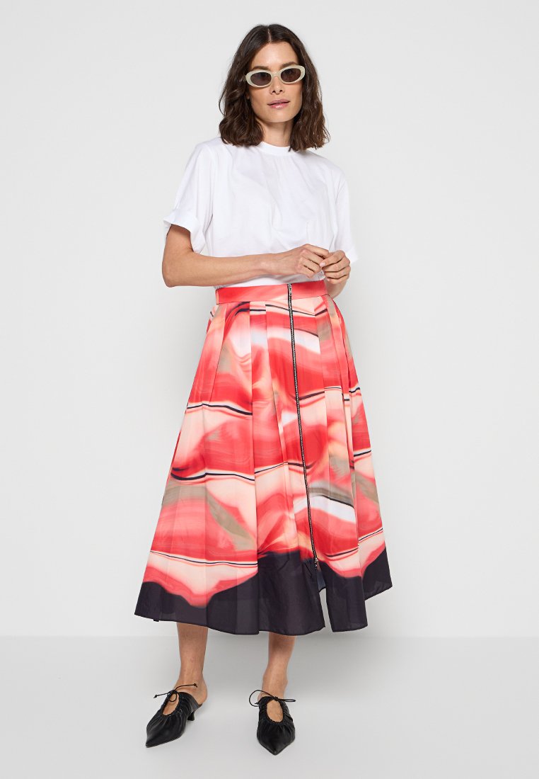 Kobieta ubrana w biały top z krótkim rękawem, midi spódnicę w abstrakcyjny wzór w czerwono-czarnych kolorach, czarne baleriny w szpic oraz białe owalne okulary przeciwsłoneczne, stojąca przy białej ścianie.