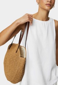 Sac en paille tressé rond avec des poignées en cuir marron, présentant une surface texturée et un sommet ouvert. Associé à un haut sans manches blanc.