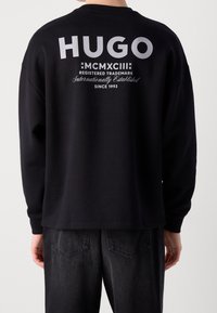 Persona che indossa una maglietta nera a maniche lunghe con la scritta "HUGO MCMXCIII Marchio registrato, riconosciuto a livello internazionale dal 1993" stampata in bianco sulla schiena.
