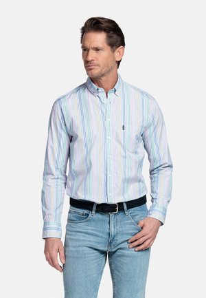 Man draagt een lang mouw, meerkleurig gestreept overhemd met knopen en een borstzak, lichtblauwe spijkerbroek en een zwarte riem, staat met handen bij zijn zakken.