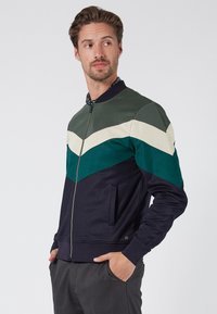 Chaqueta con cremallera que presenta un diseño de chevrón en colores verde, marino y crema. Confeccionada en una tela suave, con un cuello acanalado y bolsillos laterales.