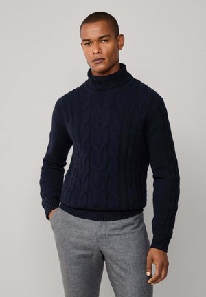 Männlicher Model trägt einen navyblauen Kabelstrick-Pullover mit Rollkragen und graue Hosen, steht mit einer Hand in der Tasche vor einem einfarbigen Hintergrund.