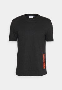 Svart bomull t-shirt med rund halsringning och korta ärmar. Har en vertikal röd Calvin Klein-logotyp tryckt på vänster sida.