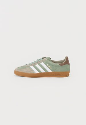 Zielony i beżowy but sportowy Adidas Gazelle z białymi paskami i gumową podeszwą, pokazany z boku na białym tle.