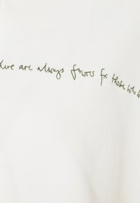 Vit t-shirt i bomull med grön, handskriven stil text broderad över framsidan, med en slät ytstruktur och avslappnad design.