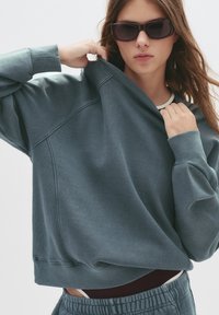 Overdimensioneret teal sweatshirt med ribbede manchetter og halsudskæring, der har en blød tekstur. Båret med matchende lyse stofbukser og lagdelt top.