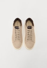 Tenisi bej din suede, cu toc din piele maro, talpă din cauciuc alb și un design clasic cu șireturi. Prezintă brandul pe limbă.