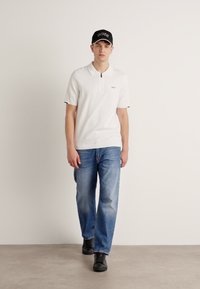 Hvit polo skjorte med korte ermer og svarte detaljer, sammen med lyseblå jeans og svarte sko. Modellen har på seg en svart caps med logo.