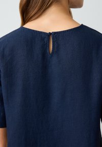 Navyfarbener Leinenoberteil mit einer Schlüsselkerbe hinten, runder Halsauschnitt und kurzen Ärmeln. Glatte Textur und minimalistisches Design.