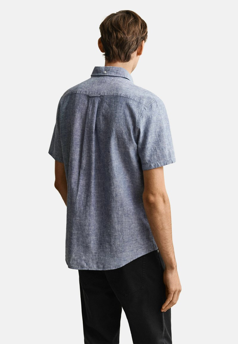 Uomo che indossa una camicia chambray a maniche corte color celeste e pantaloni neri, visto di spalle su uno sfondo semplice.