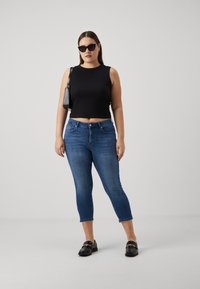 Top corto negro, jeans ajustados de mezclilla azul, mocasines negros con detalle de cadena y gafas de sol negras, todo sobre un fondo liso.
